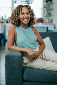 An image of Angel Whaley, a lighter-skin Black woman with curly hair wearing a pale blue shirt and white pants, sitting and smiling into the camera. 